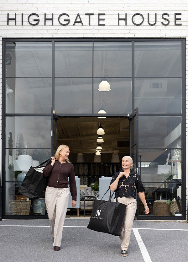 Two women walk out of a storefront labeled HIGHGATE HOUSE, each carrying a large black tote bag with a white monogram logo. The scene is set in front of a modern glass-front building with a white brick facade.