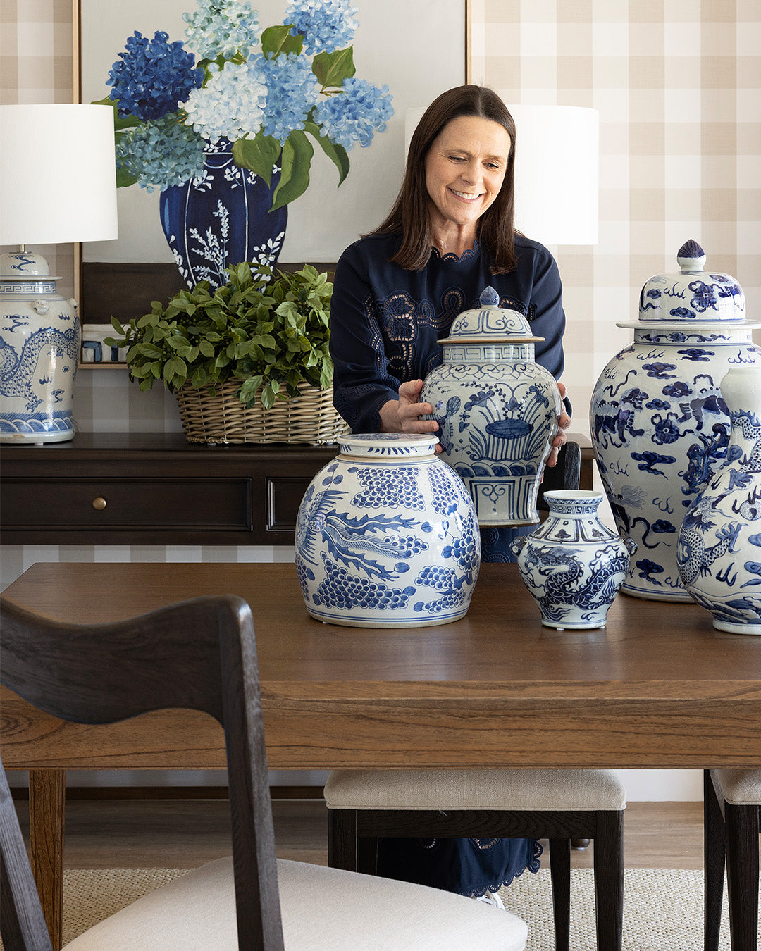 A woman stands behind a wooden table displaying a collection of blue and white porcelain garden jars and vases, with a matching blue and white floral vase and lamps in the background.