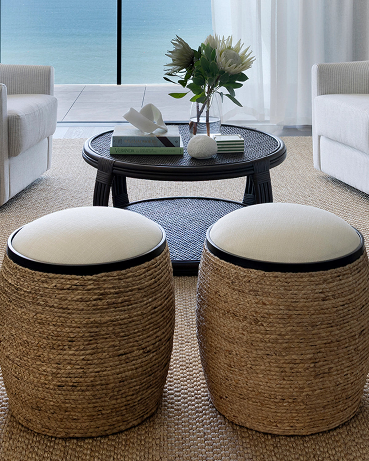 Two round woven rope ottomans with cream cushions in a modern living room setting, positioned in front of a black coffee table and overlooking a sea view.
