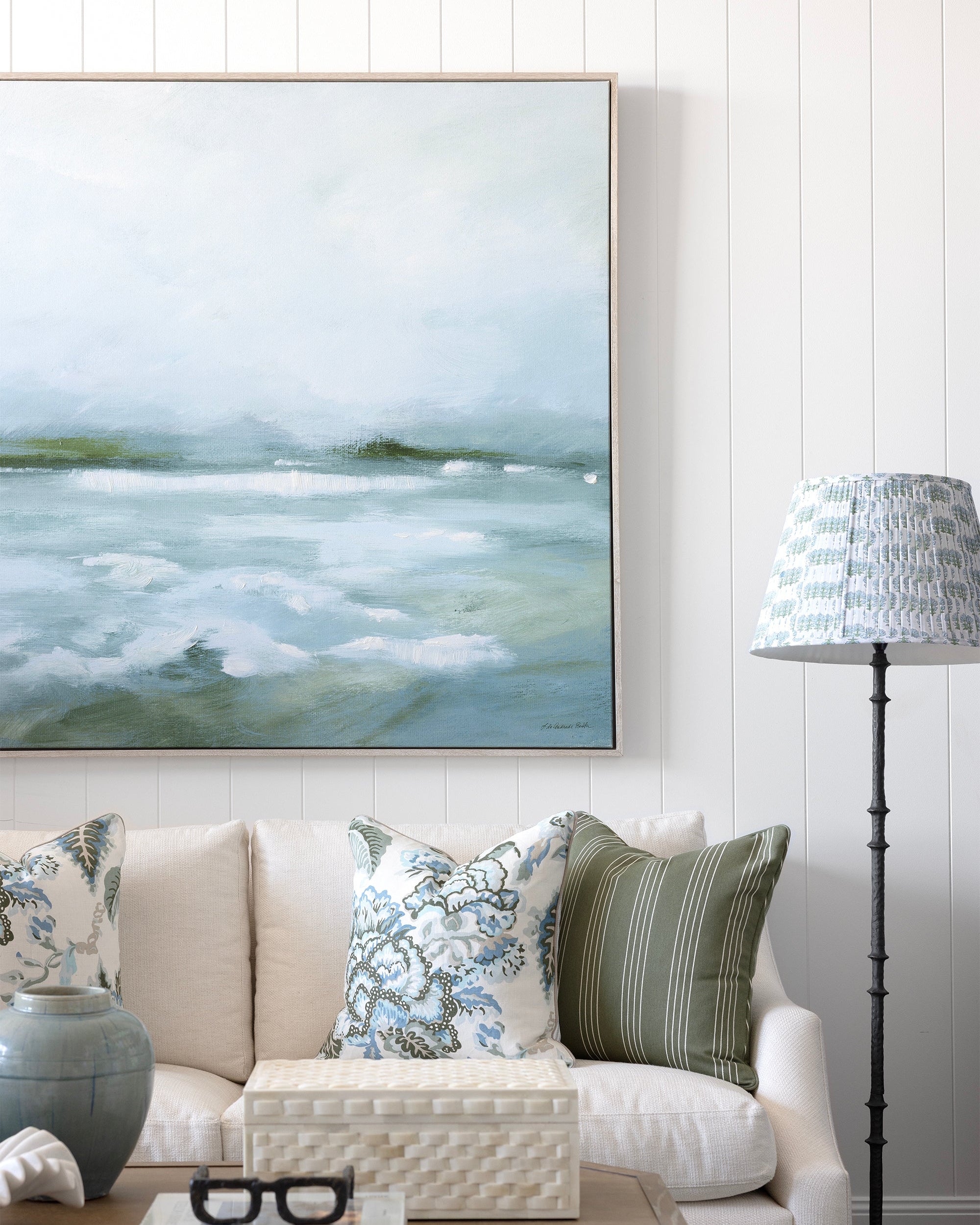 A large framed landscape painting of misty waves hangs on a white paneled wall above a cream sofa with patterned throw pillows. A wooden coffee table, a blue-gray ceramic vase, and a black floor lamp with a patterned shade are visible in the styled room.