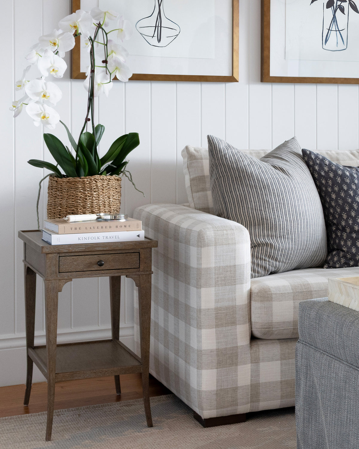A styled living room corner featuring a beige and white plaid sofa with striped and patterned accent pillows, a rustic brown wooden side table holding a potted white orchid and books, and white paneled walls with framed art.
