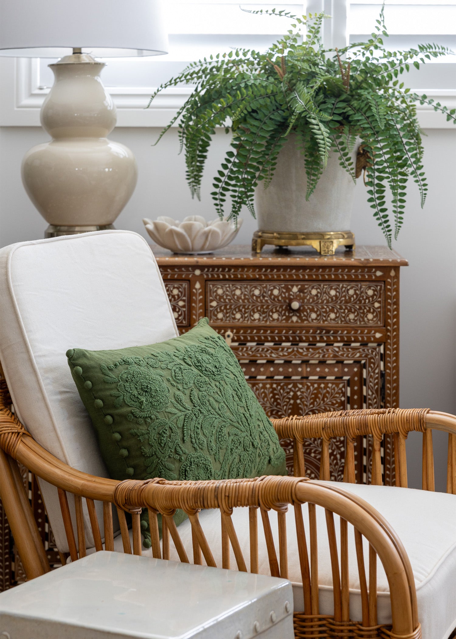 Mara cabinet styled in a living space. With a Rattan chair in front. The cabinet is styled with a large faux fern arrangement a small marble lotus bowl and a cream lamp.