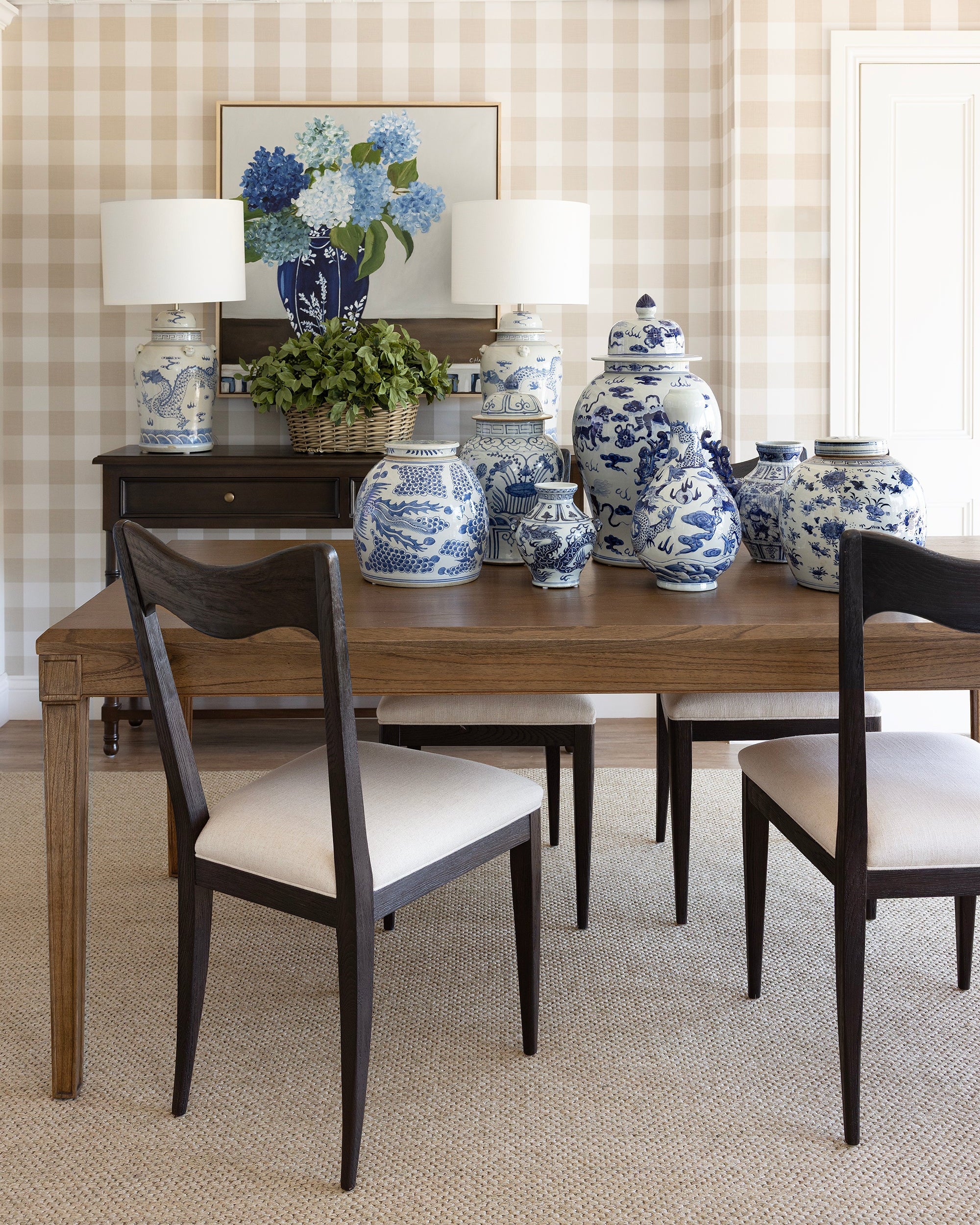 A styled dining room scene featuring a wooden dining table with four dark wood chairs that have cream upholstered seats, set against a beige gingham checkered wall. On the table are several blue and white porcelain ginger jars, and on the dark wood sideboard behind it are two matching blue and white table lamps and a framed floral print.