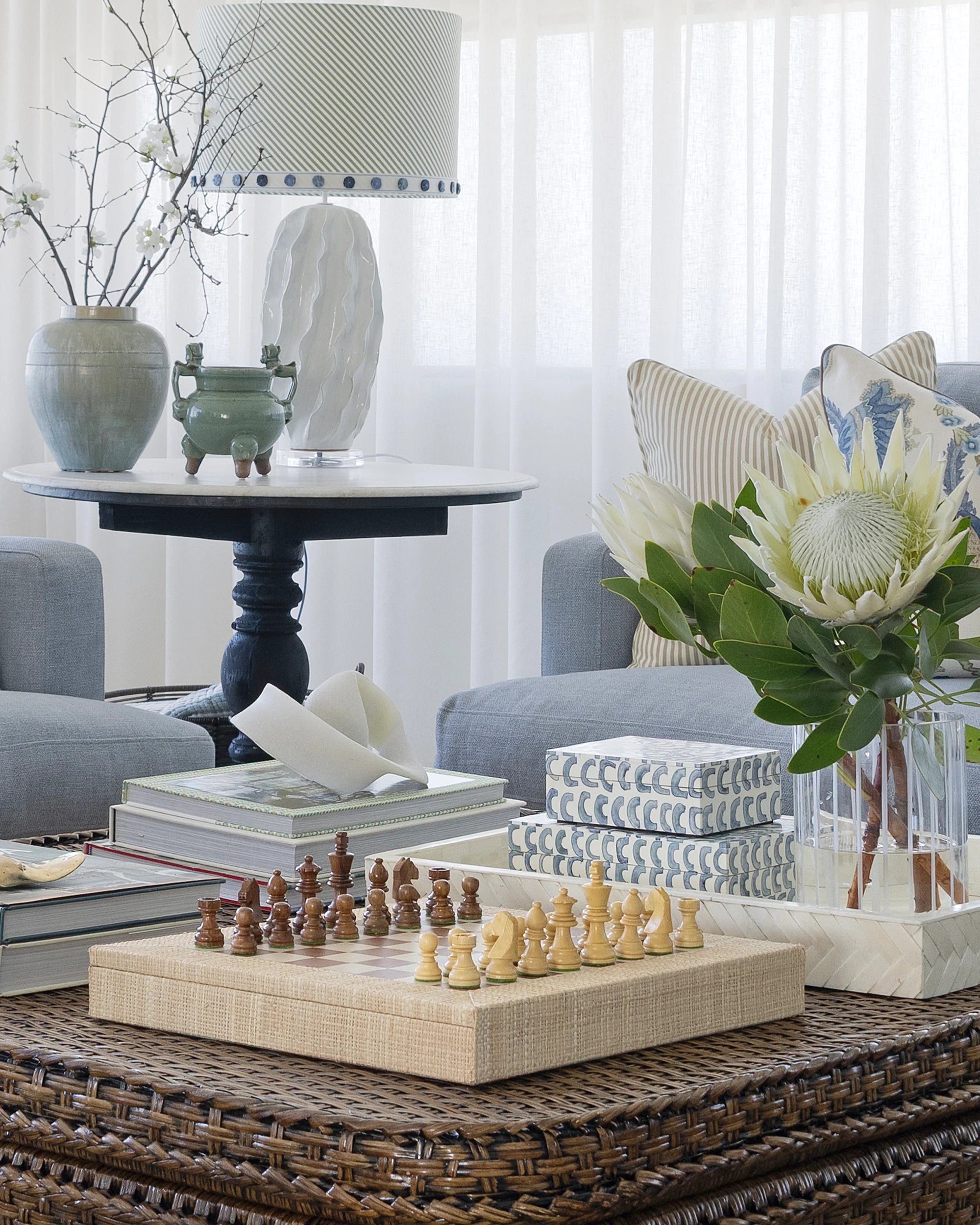 A styled living room scene showing a woven coffee table with a wooden chess set in the foreground, books, decorative boxes, a vase of protea flowers, a gray sofa, and a side table with a lamp and ceramic vases in the background.