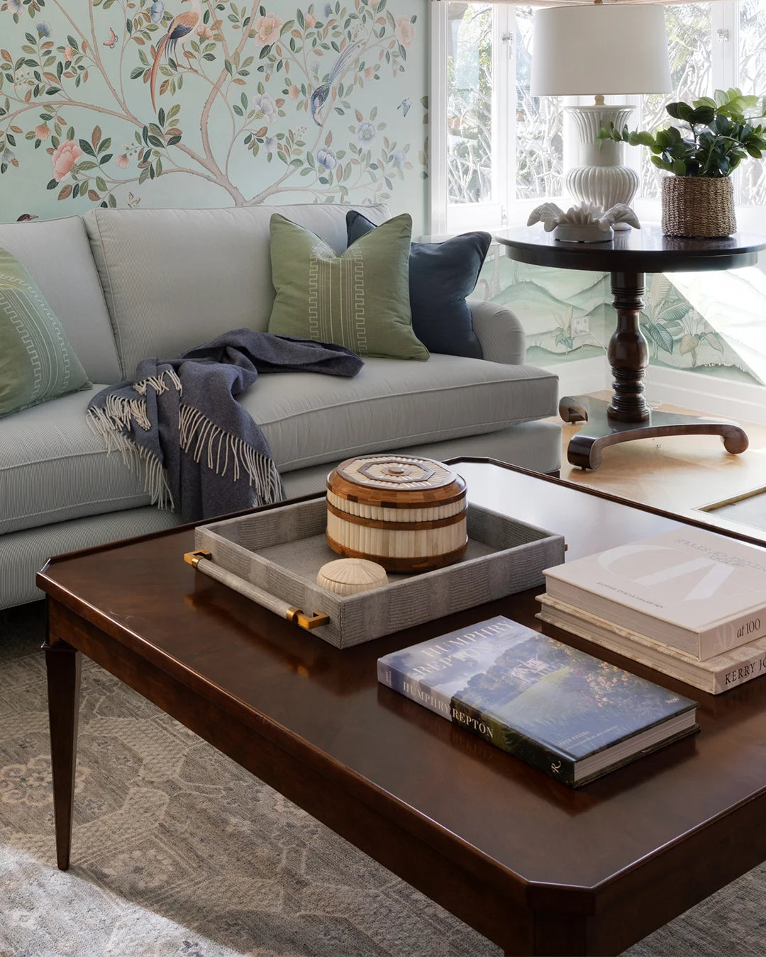 Square coffee table styled with books, and a tray and box, grey sofa with blue and green cushions behind. Botanical wallpaper with centre table to the side of sofa.