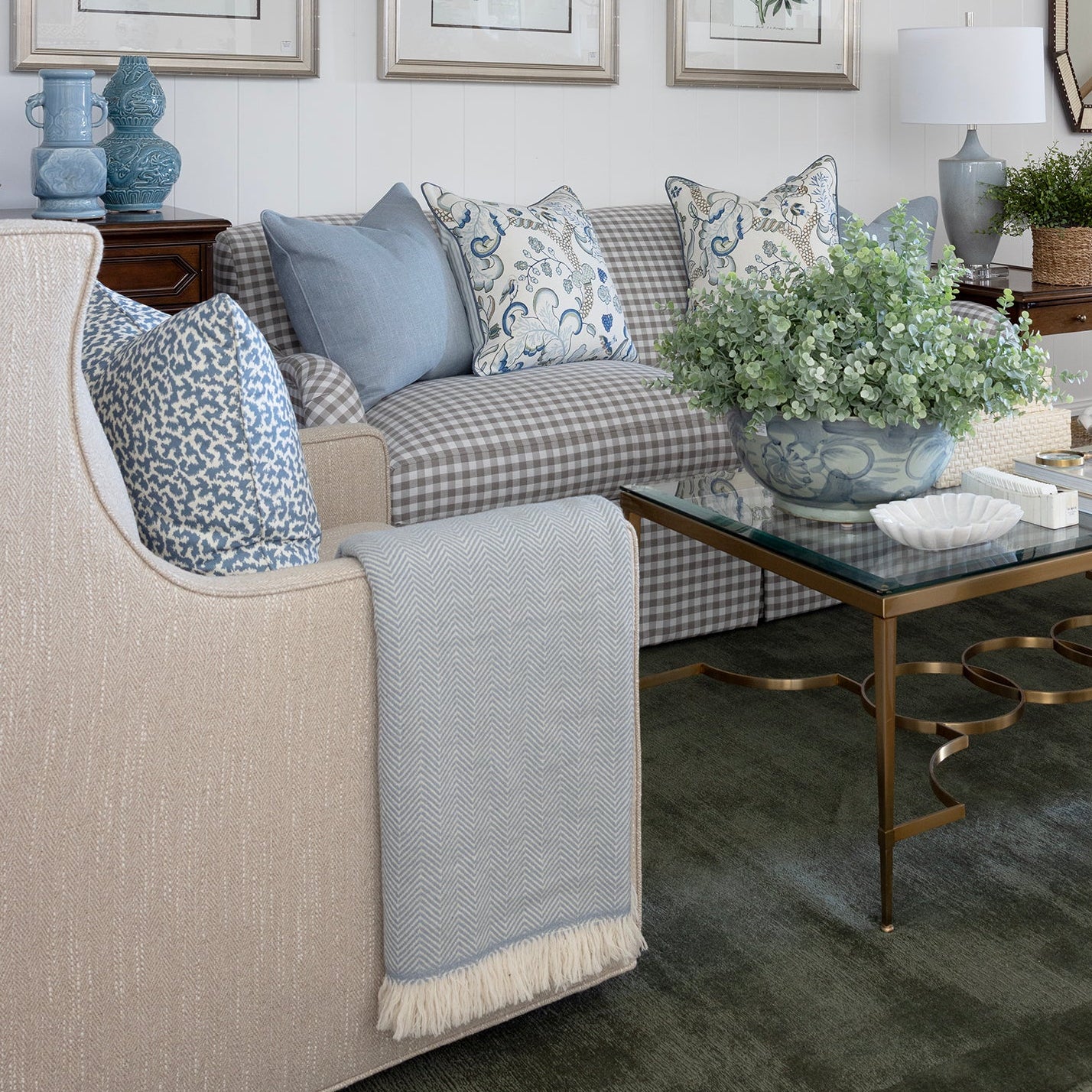 A styled living room interior featuring a beige armchair, a blue-and-white checkered sofa with blue accent pillows, a glass-top coffee table with gold legs, framed botanical prints on the wall, and a white ceiling with exposed beams.