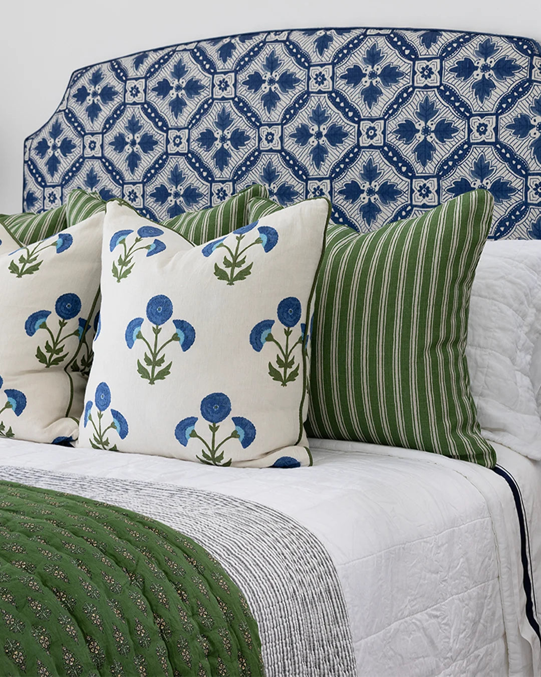 A styled bed with a blue and white geometric patterned headboard, white bedding, a green quilt, and decorative throw pillows including one with blue flowers on cream and another with green and white vertical stripes.