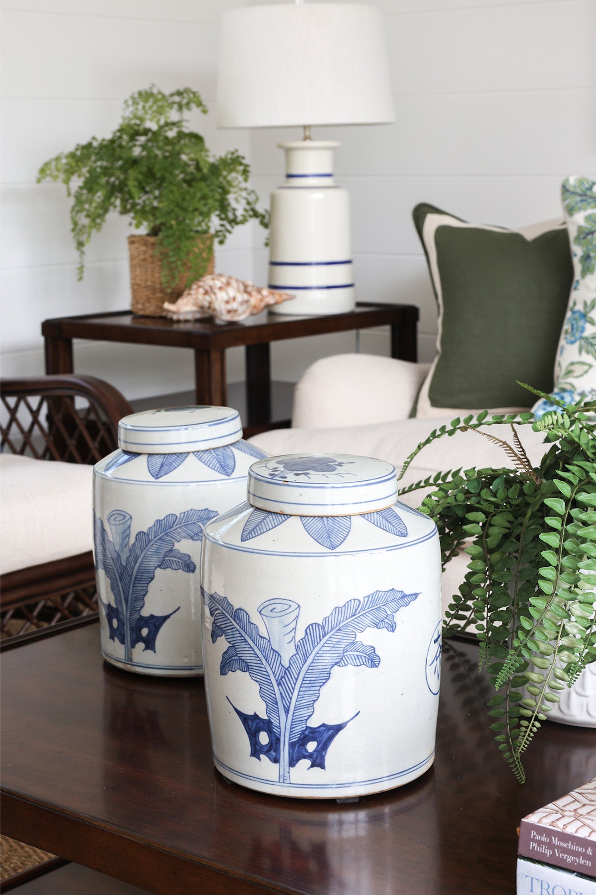 Two blue and white ceramic jars on a wooden table with a lamp and plants in the background.
