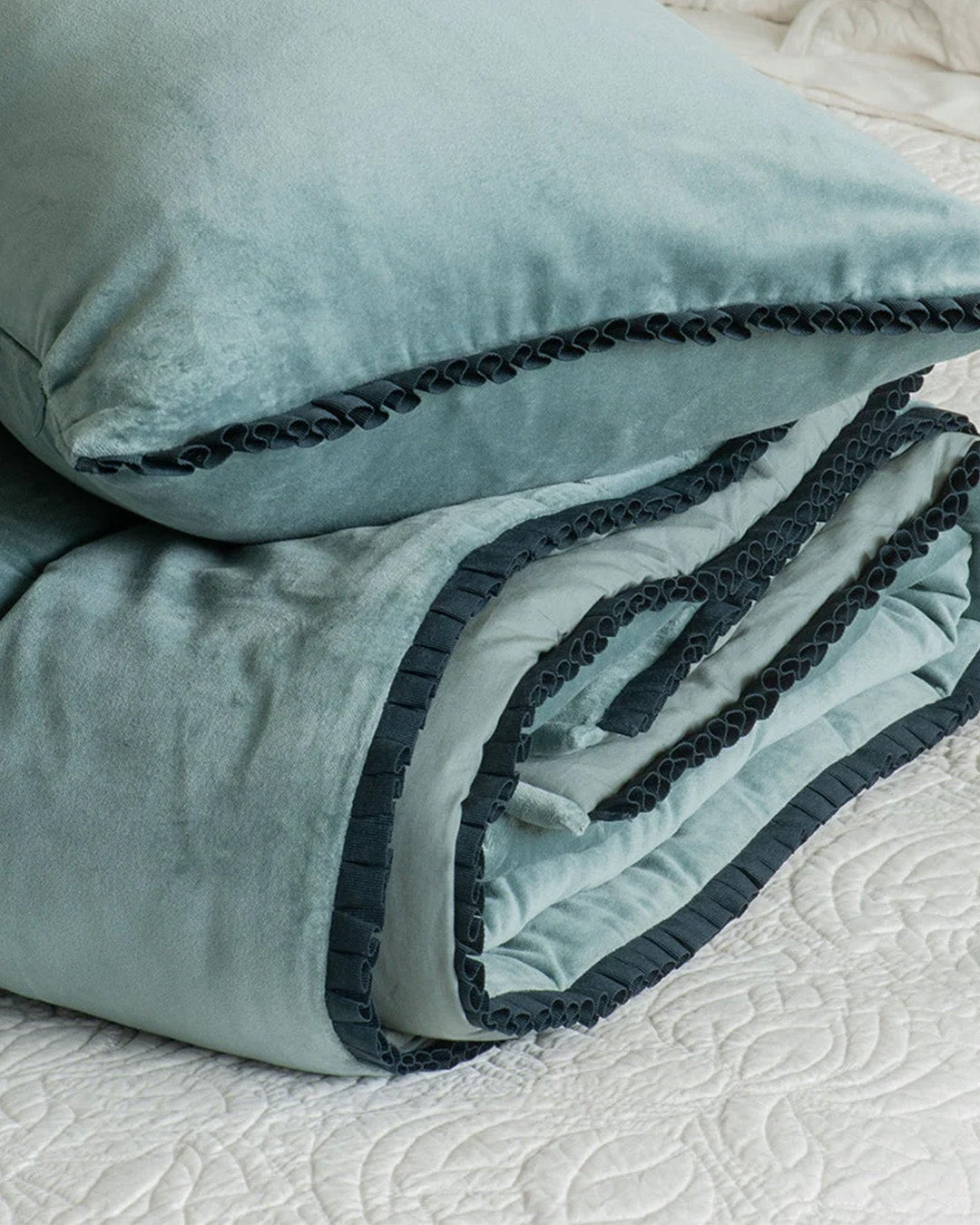 Folded blue velvet comforter, with a dark blue green ruffled edge, sitting on white embroidered quilt, matching pillow sitting on top.