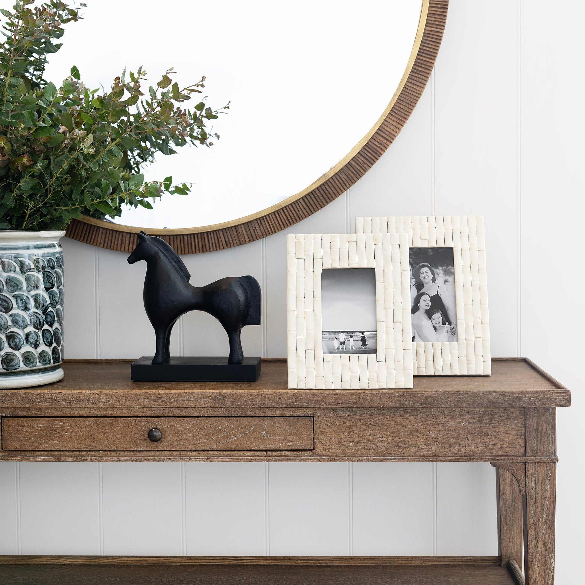 Wooden console table with decorative items including a horse statue, plant, and framed pictures against a white wall.