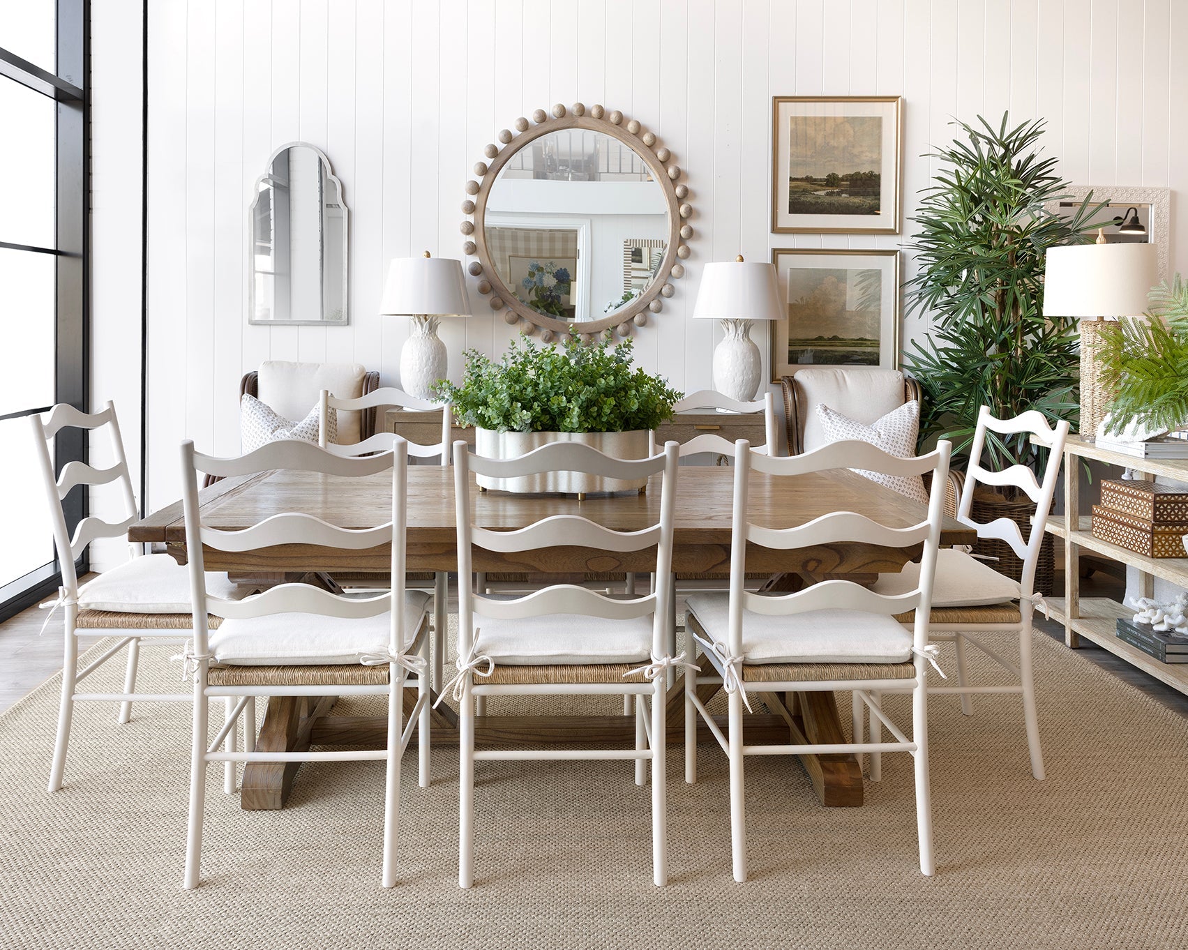 A dining room scene featuring a rustic wooden dining table surrounded by six white ladder-back chairs with white upholstered seats. The room is styled with a large round decorative mirror, white table lamps, framed art, potted green plants, and a white sideboard on a light beige rug.