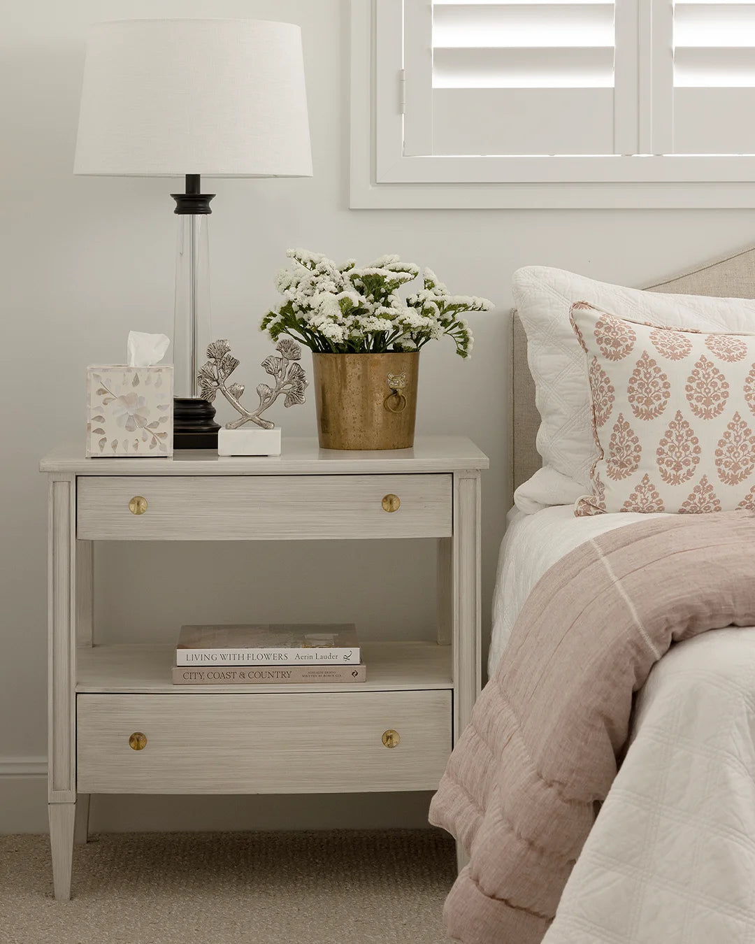 Soft grey bedside table styled with lamp, tissue box, metal vase of flowers and silver branch decorative object. Sitting beside bed with pink and white bedding.