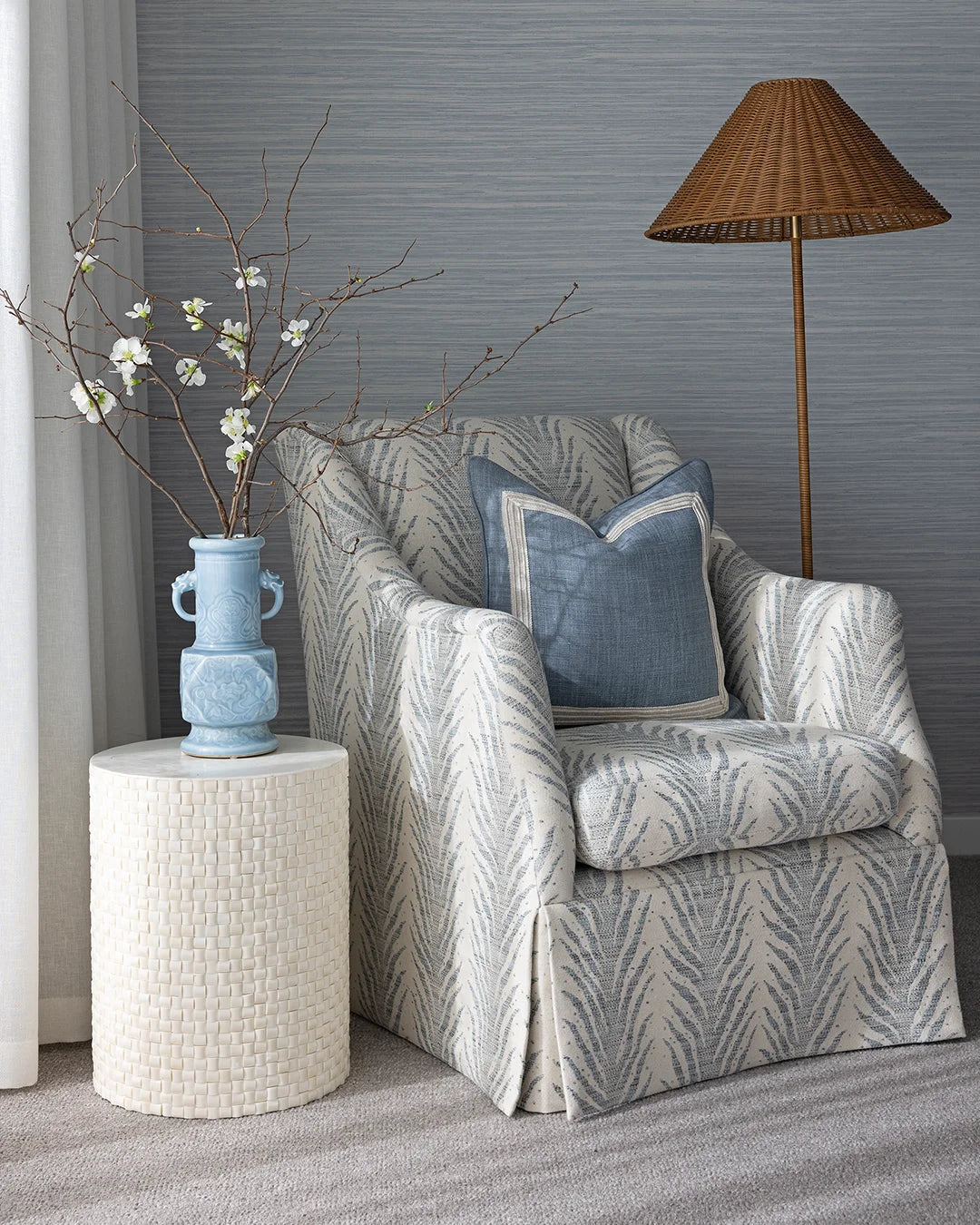 A classic slipcovered armchair with a gray and white leaf pattern, styled with a blue accent pillow, next to a textured white side table with a blue vase, against a gray textured wall.