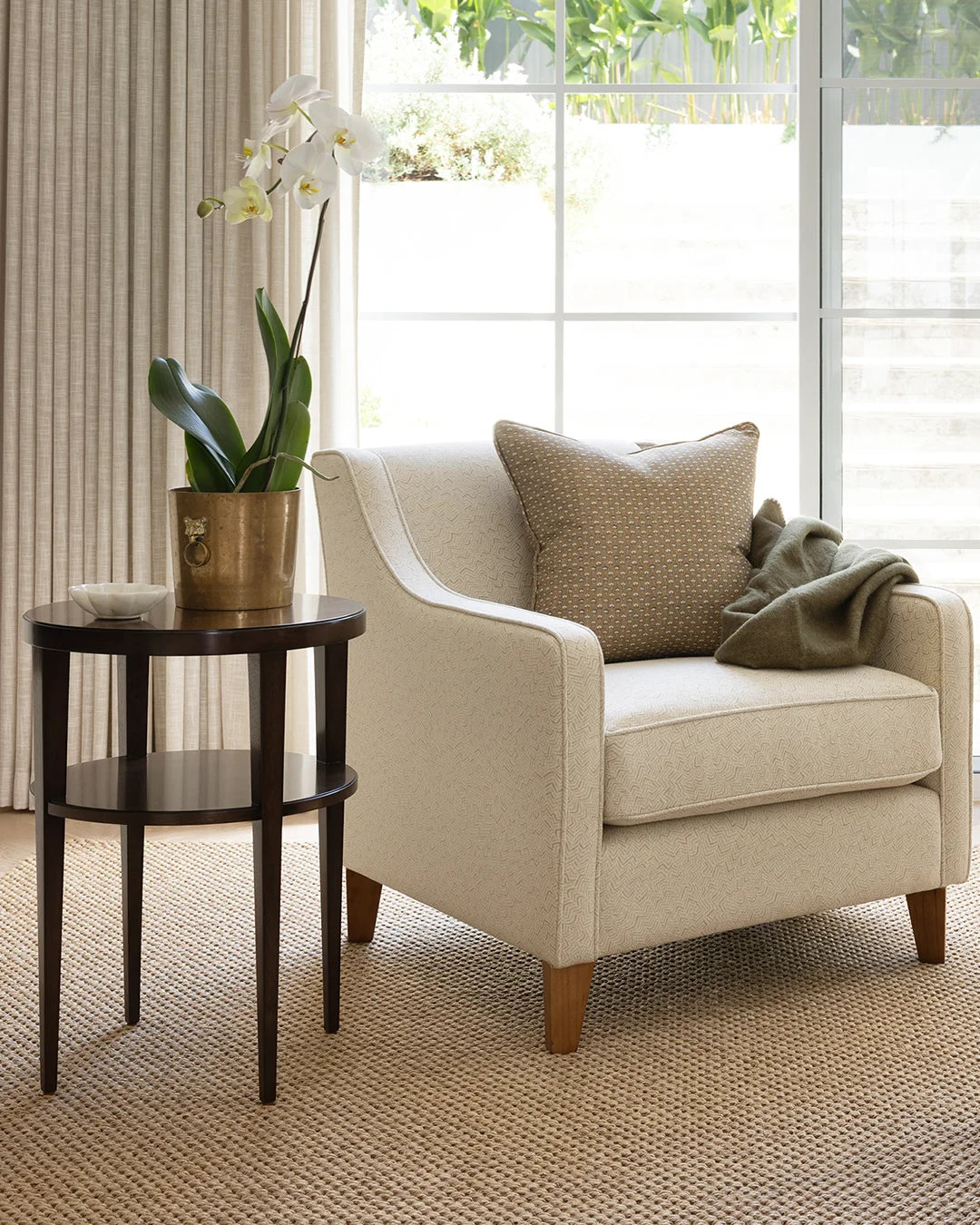 A cream upholstered armchair in a bright living room setting, styled with a beige patterned throw pillow, a folded olive green blanket, and a dark wood side table holding a potted white orchid.