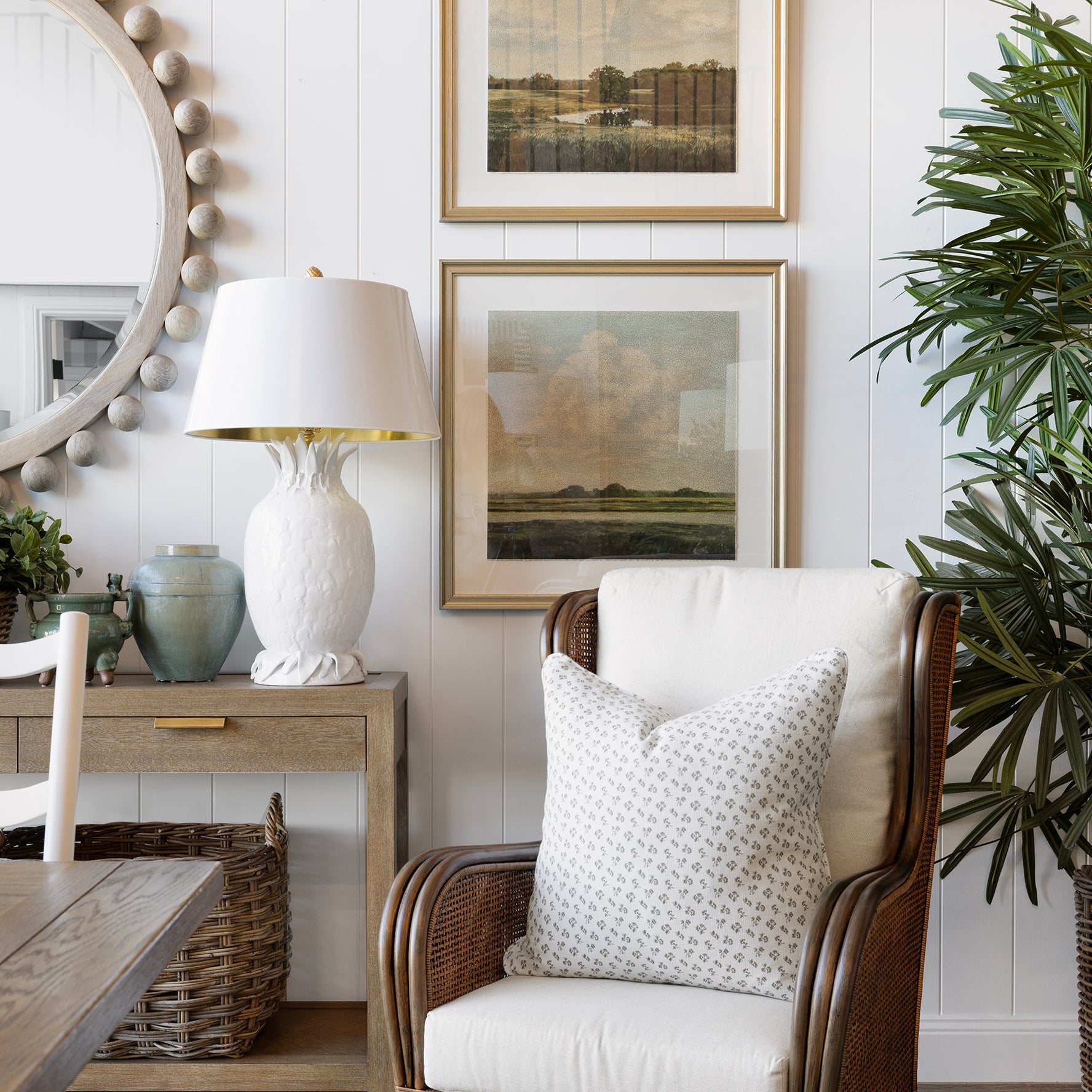 A styled interior scene featuring a brown wicker armchair with white cushions, a white table lamp, a wooden console table, a round beaded mirror, and two framed landscape prints on a white shiplap wall.