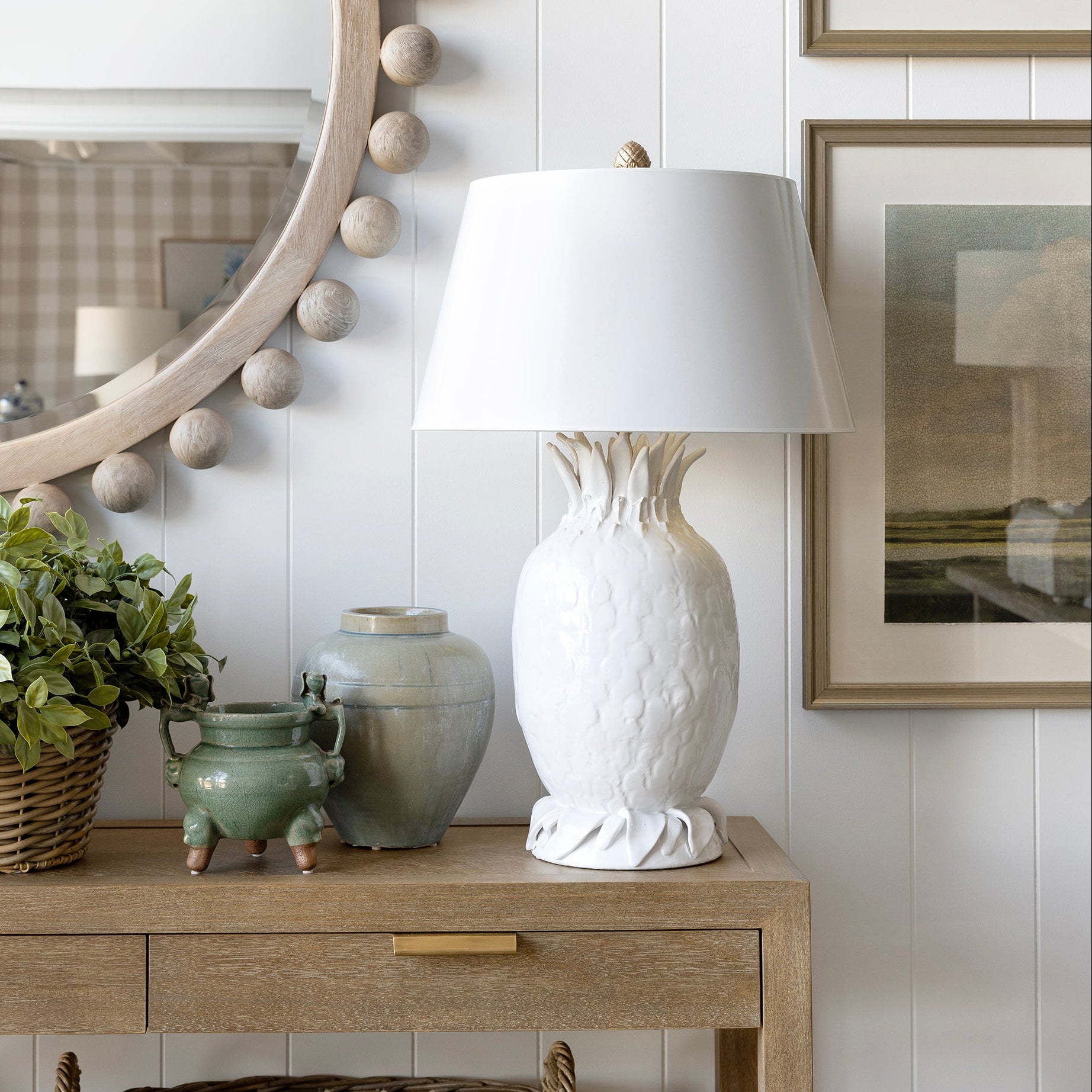 A white textured ceramic table lamp with a tapered shade sits on a light wood console table against a white shiplap wall, styled with a green plant, ceramic vases, a round beaded mirror, and framed landscape prints.